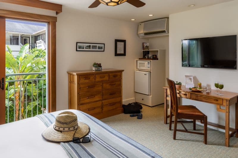 A hotel room interior featuring a king bed, a wooden dresser and a matching desk with a chair, a wall-mounted TV, and an open sliding glass door that leads to a balcony with lush green foliage.