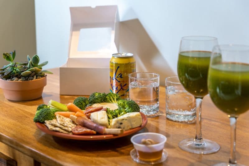 A close-up of a wooden table featuring two wine glasses filled with green juice, a platter of broccoli, crackers, and deli meats, a small container of dip, and a succulent in a terracotta pot.