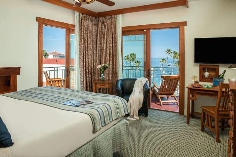 A spacious hotel bedroom featuring a queen-sized bed with a blue-and-white striped throw, a flat-screen TV on the wall, and two separate sliding glass doors that lead to private balconies overlooking the ocean and harbor.