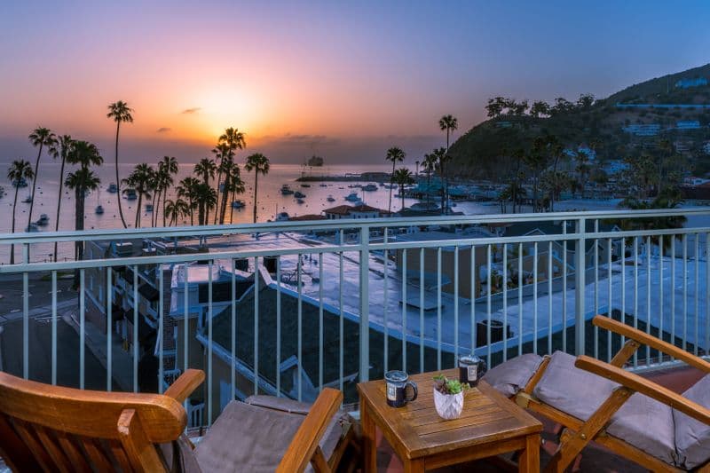A private hotel balcony at dusk, featuring two wooden lounge chairs and a small table overlooking a calm harbor at sunset, with silhouetted palm trees and a soft orange glow on the horizon.