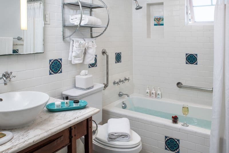 A modern hotel bathroom featuring white subway tile walls accented with blue and green floral tiles, a marble-topped vanity with a white vessel sink, and a bathtub with integrated safety handrails.