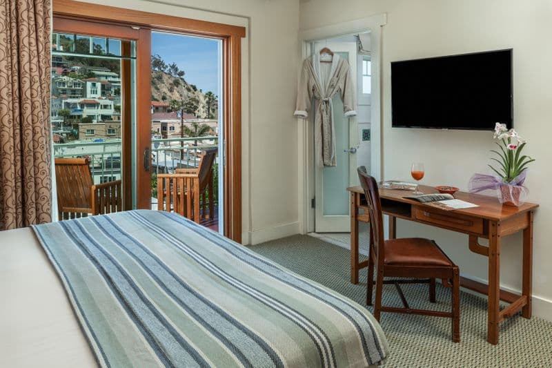 A bright hotel room with a wooden writing desk and chair, a wall-mounted TV, and an open sliding door revealing a balcony with wooden furniture and a view of a hillside town.