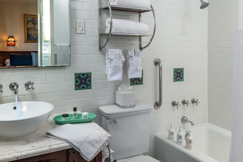 A bright, white-tiled bathroom featuring a marble vanity with a vessel sink and Fiesta ware accessories, a combination bathtub and shower with silver fixtures, and decorative green and blue patterned Catalina tiles.