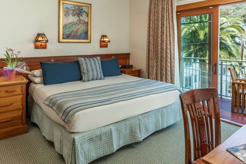 A well-lit bedroom featuring a large bed with a light blue bed skirt and a blue and white striped throw, a wooden dresser with a small plant, and a sliding glass door that opens to a balcony with a view of a lush palm tree.