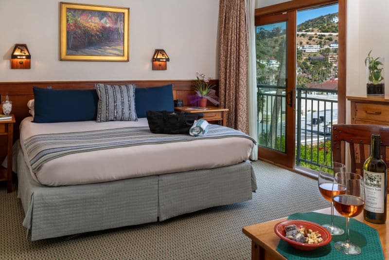 A cozy hotel bedroom with a king bed featuring a blue and white striped throw, a wooden headboard, and two wine glasses and a snack plate in the foreground, with an open sliding door revealing a hillside view.