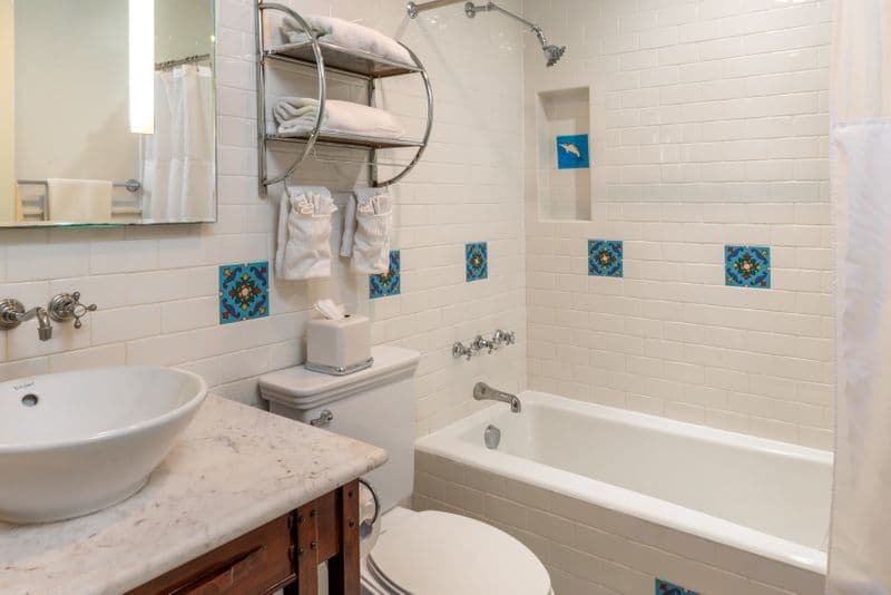 A clean, white-tiled hotel bathroom featuring a marble vanity with a vessel sink, a combination bathtub and shower, and decorative blue and green floral Catalina tiles accented along the walls.