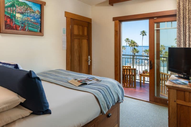 A bright hotel room with a queen-sized bed featuring a blue-and-white striped throw, a wooden dresser with wine glasses in the foreground, and a sliding glass door leading to a private balcony with a view of a scenic harbor and palm trees.