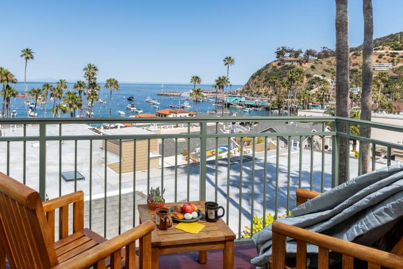 A private hotel balcony featuring two wooden armchairs and a small table set with a snack plate and a black mug, looking out over a sun-drenched harbor filled with boats and lined with tall palm trees.