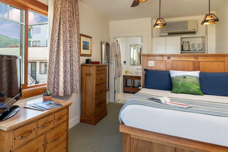 A bright hotel room featuring a bed with a blue-and-white striped throw, a wooden dresser with brochures and wine glasses in the foreground, and a window offering a view of neighboring buildings and green trees.