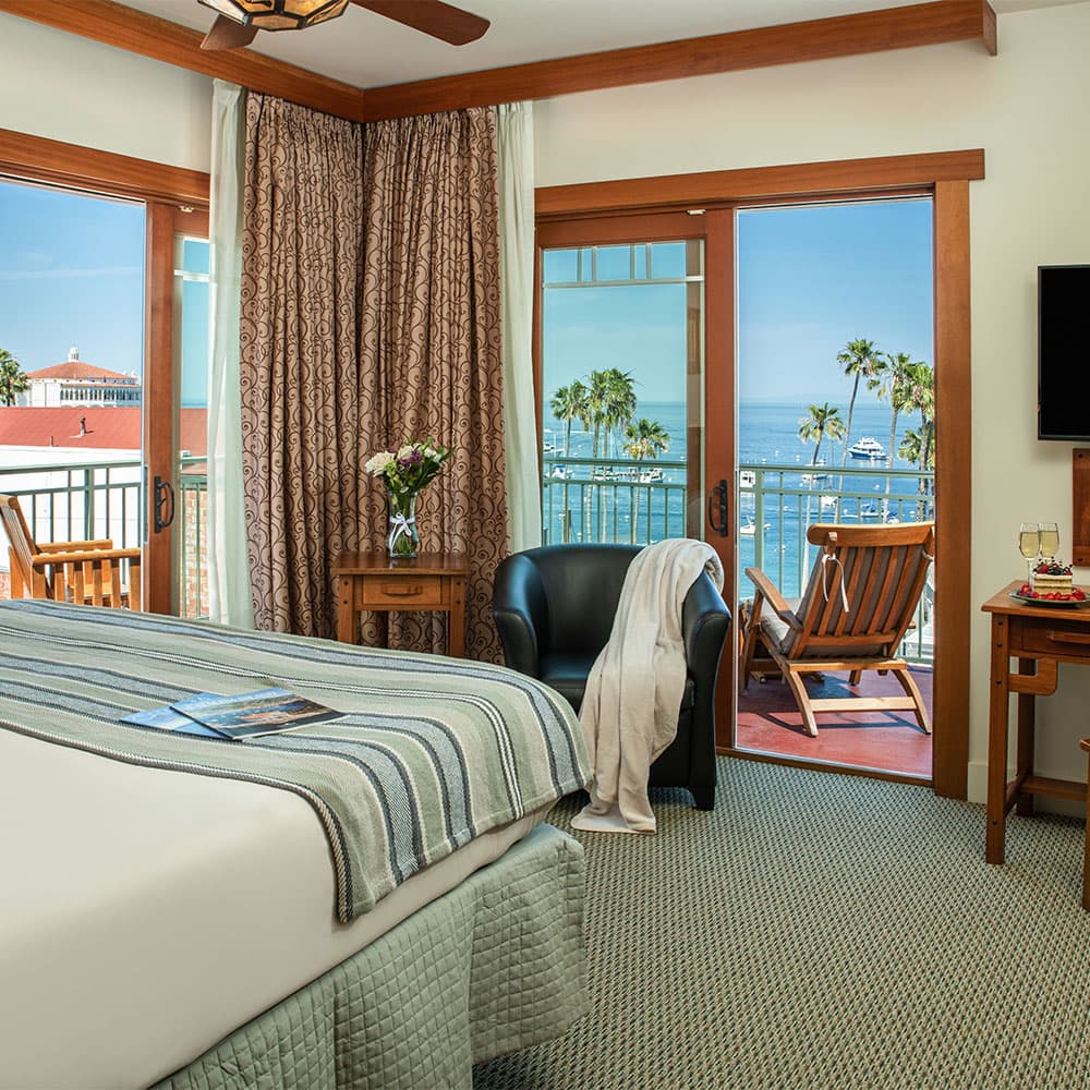 Cozy hotel room with a bed, chairs, and a balcony view of the ocean and palm trees.
