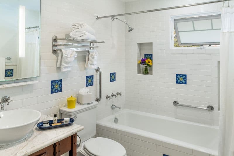 A clean, white-tiled hotel bathroom featuring a marble vanity with a vessel sink, a combination bathtub and shower with silver fixtures, and decorative blue and green Catalina tiles.