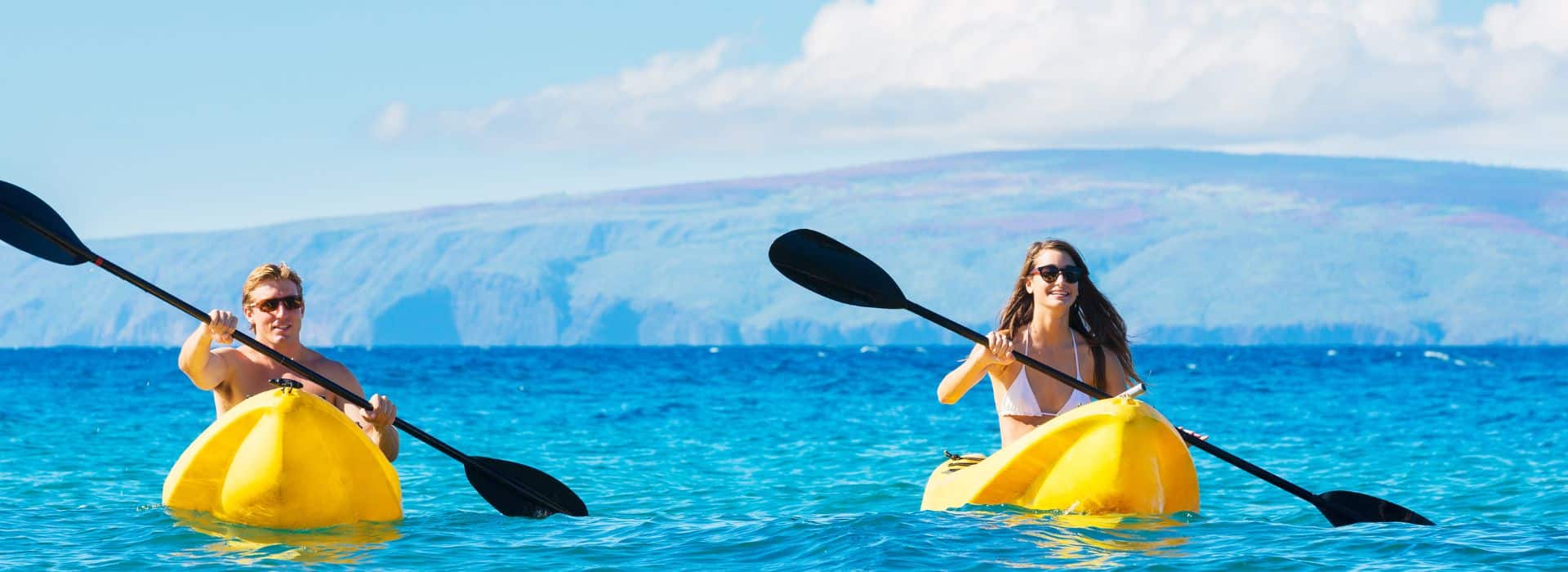 Two people paddle yellow kayaks in a clear blue ocean with a mountainous background. Two people paddle yellow kayaks in a clear blue ocean with a mountainous background.