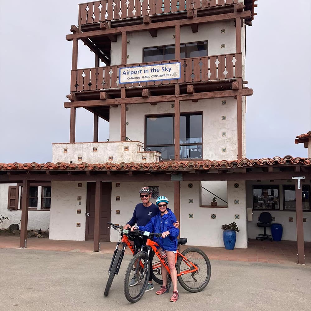 A man and a woman stand with bicycles in front of a building labeled "Airport in the Sky" under an overcast sky.