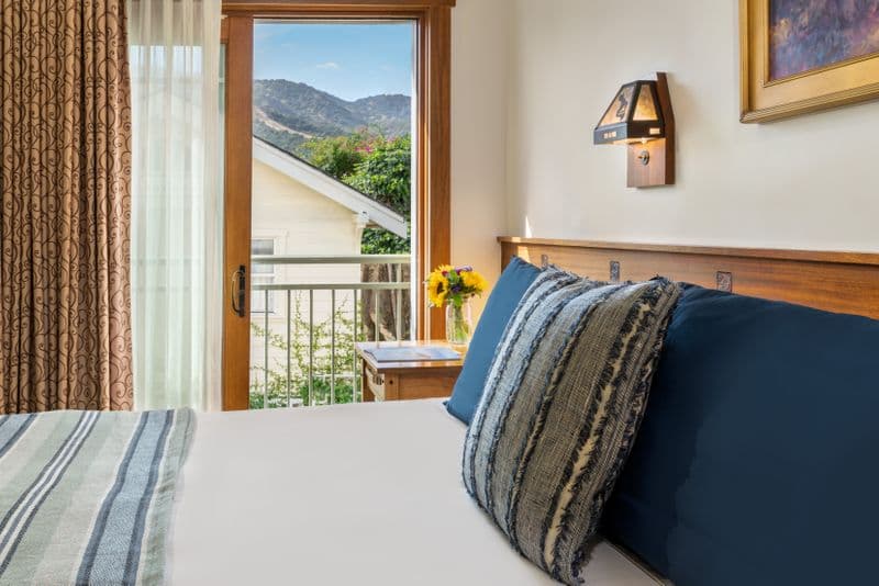 A well-lit hotel bedroom featuring a king-sized bed with a blue and white striped throw, a wooden headboard, and a large sliding glass door that offers a clear view of a lush green hillside.