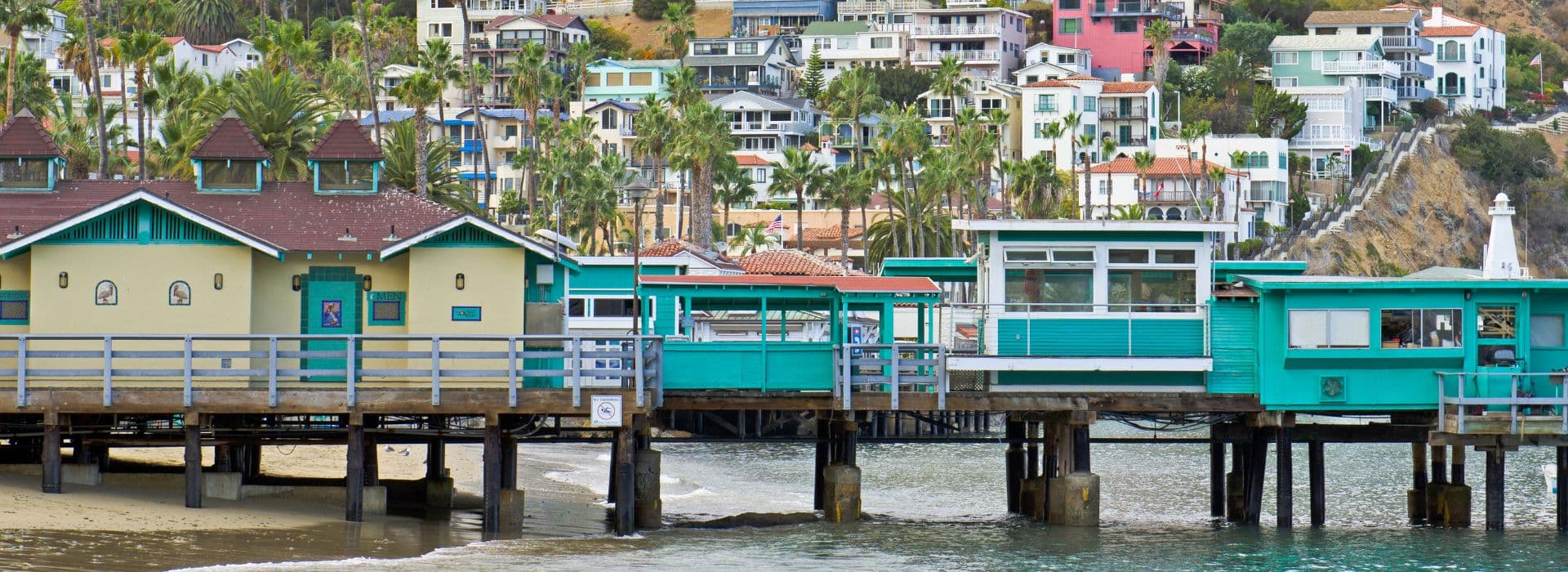 A colorful pier extends over calm water, surrounded by palm trees and vibrant hillside homes.