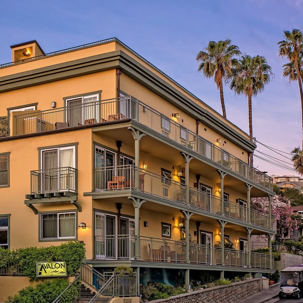 A three-story yellow building with balconies, surrounded by palm trees.