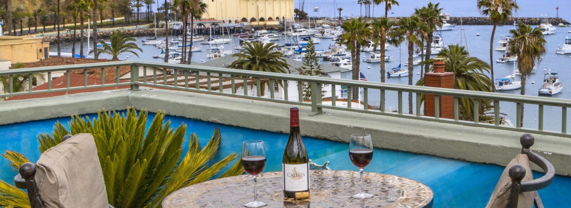 A scenic view overlooking a marina with palm trees, featuring a table with a bottle of wine and two glasses.