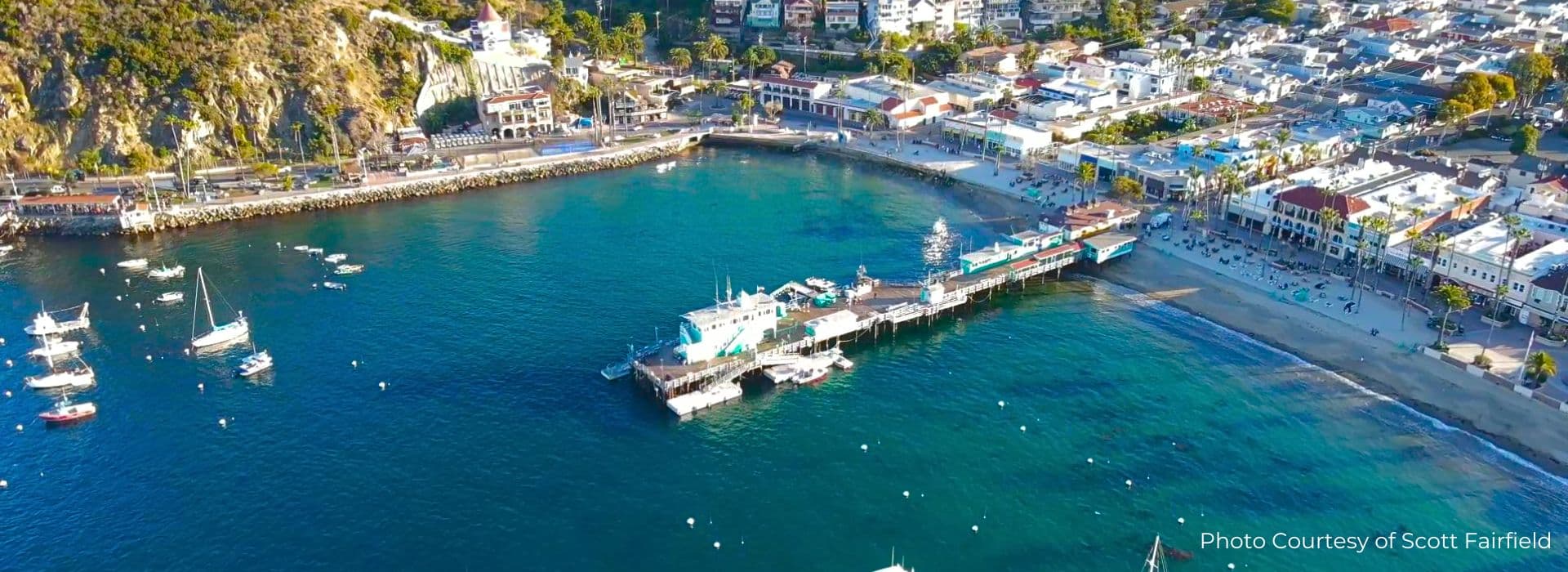 Aerial view of a coastal harbor with boats, a pier, and colorful buildings along the shore.