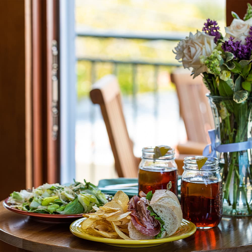 A sandwich, salad, and chips on a table beside two glasses of iced tea and a flower vase.