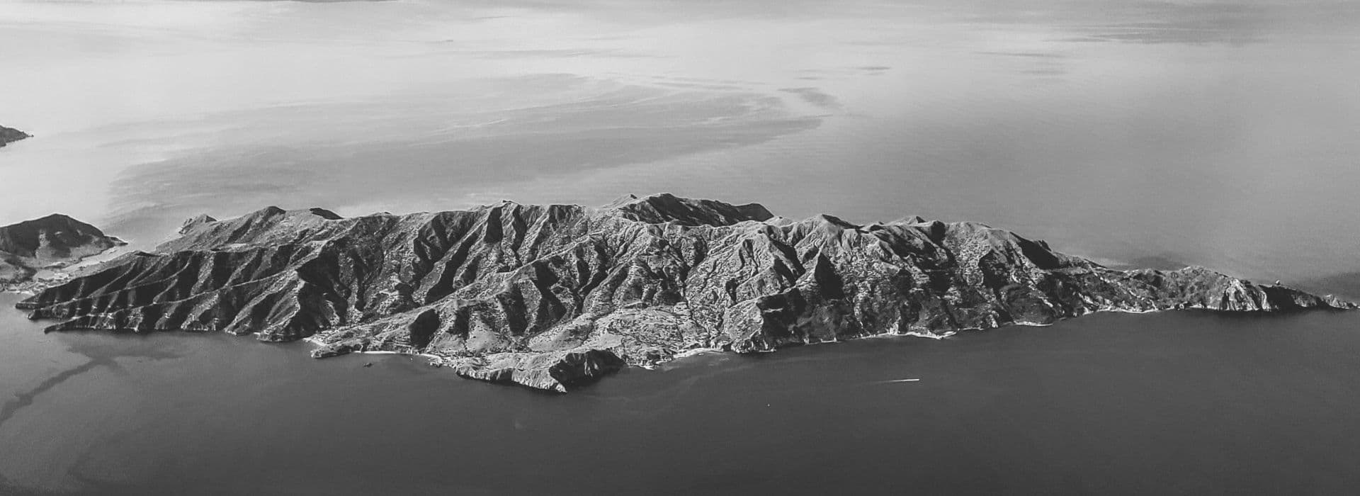 Aerial view of a rugged, mountainous island surrounded by water, presented in black and white.