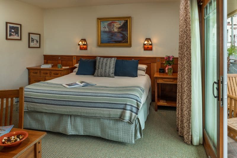 A bright hotel bedroom featuring a bed with navy blue and striped pillows, a wooden nightstand with a small vase of flowers, and an open sliding glass door leading to a balcony with a view of a palm tree.