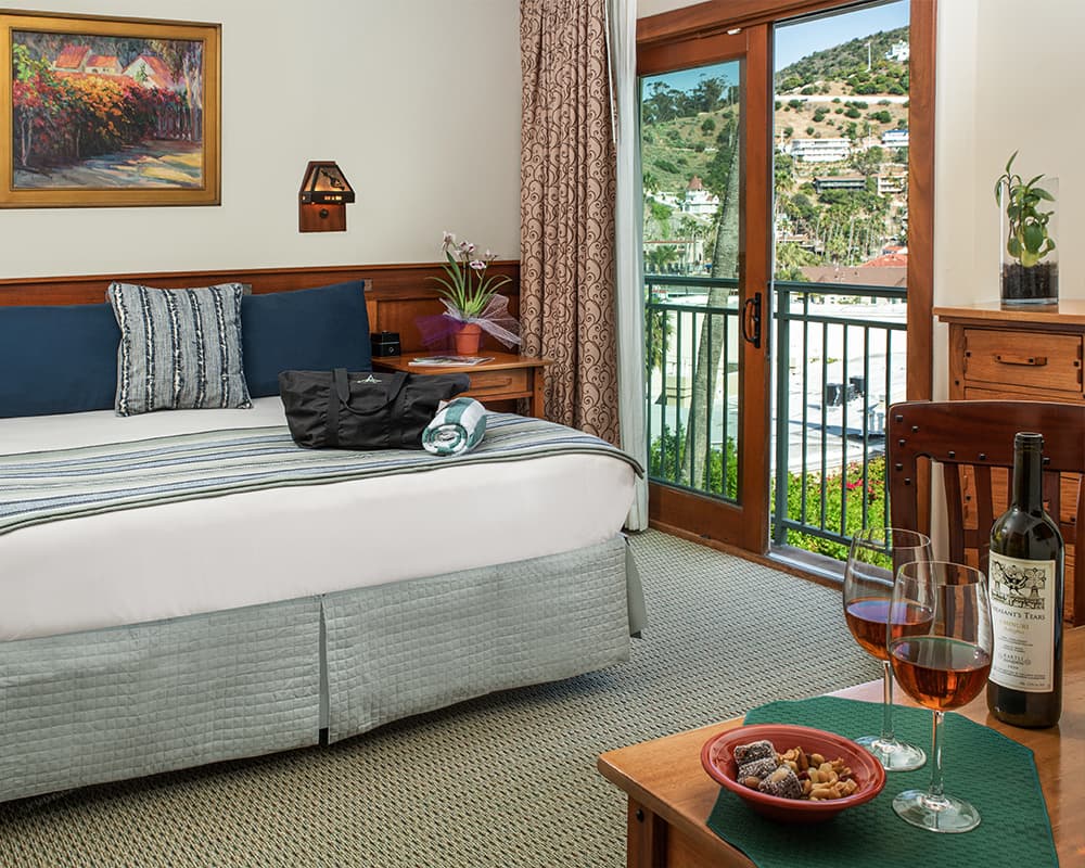 A cozy hotel room with a queen bed, balcony view, and a table set with wine and snacks.