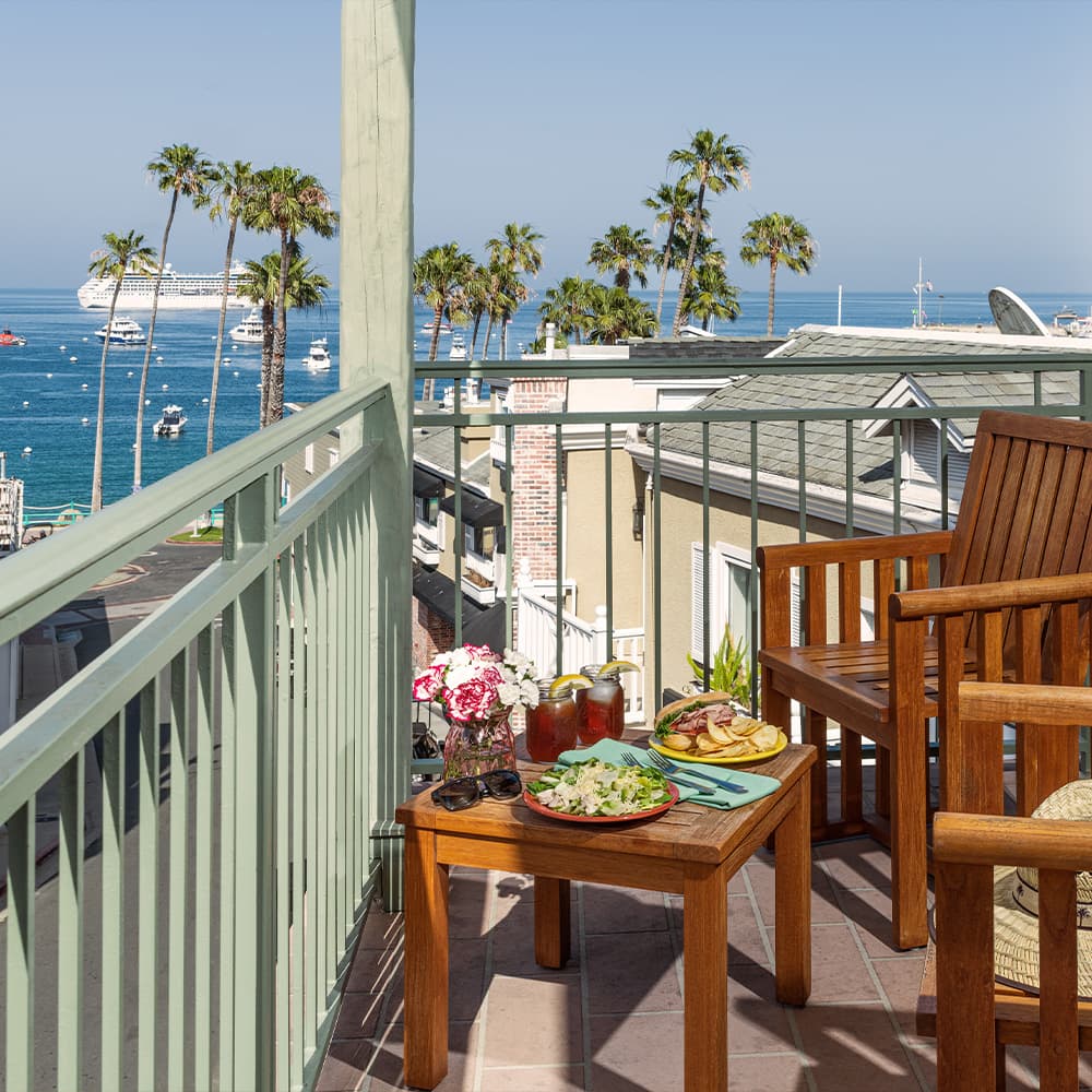 A balcony with wooden chairs overlooks the ocean, featuring a table set with food and drinks, and surrounded by palm trees.