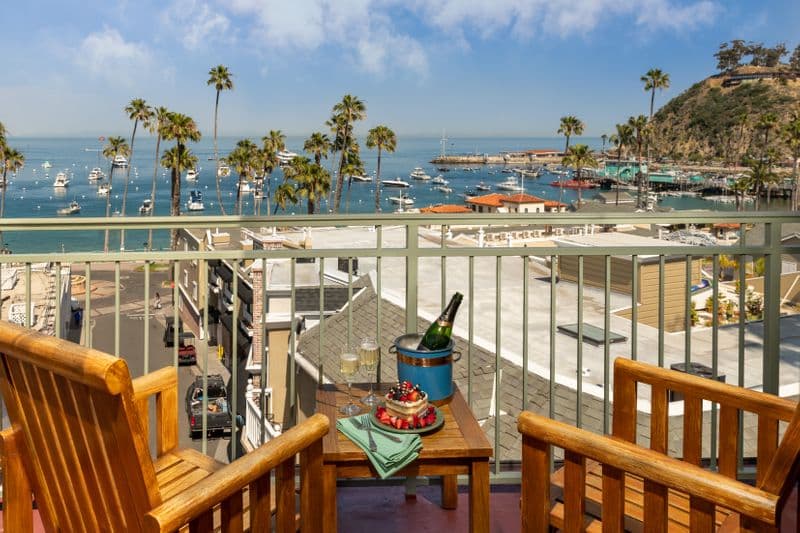 A sunny hotel balcony featuring two wooden armchairs and a small table set with a bottle of champagne in an ice bucket and a plate of snacks, overlooking a harbor filled with boats and lined with palm trees.