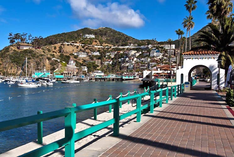 A scenic waterfront promenade with turquoise railings, boats, and hillside homes under a blue sky.