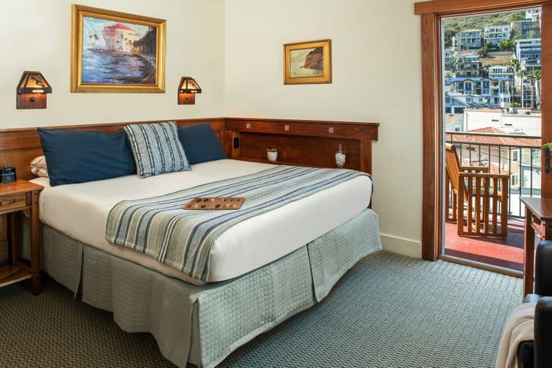 A bright hotel room featuring a queen bed with a blue-and-white striped throw, a wooden headboard with built-in lamps, and a view of a scenic coastal hillside through an open sliding glass door.