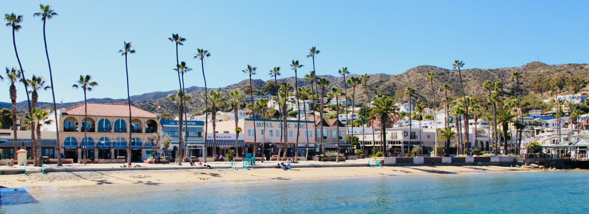 Beach front full of retail and lodging businesses surrounded by palm trees and hills in the background