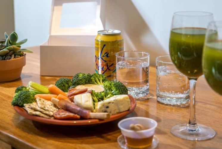 Close up view of plate with cheese, meat, crackers, veggies, and dip next to drinks and small plant on wooden desk