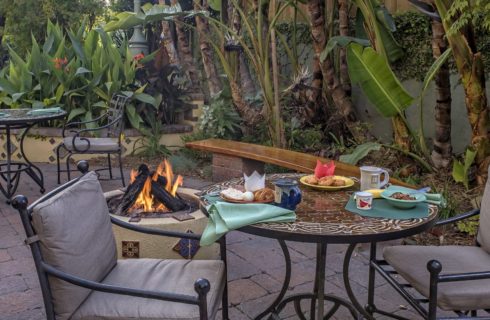 Stone patio with metal patio tables, chairs with gray cushions, and firepit surrounded by lush gardens