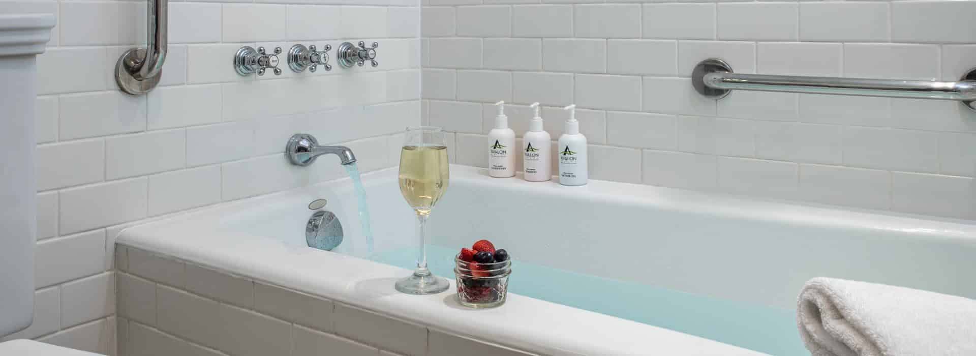Close up view of bathtub surrounded by white tiles, glass of white wine, jar of fresh fruit, and branded toiletries