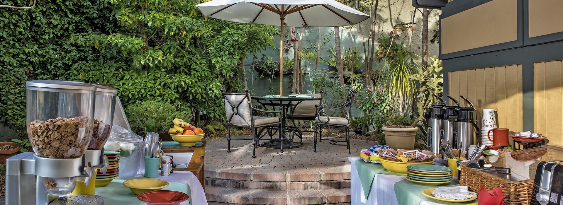 Stone patio with metal patio tables, chairs with gray cushions, patio umbrella, lush gardens and tables set up with continental breakfast items