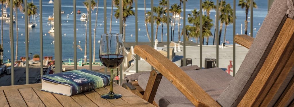Close up view of glass with red wine and book on wooden table and wooden patio chair with view of the harbor in the background